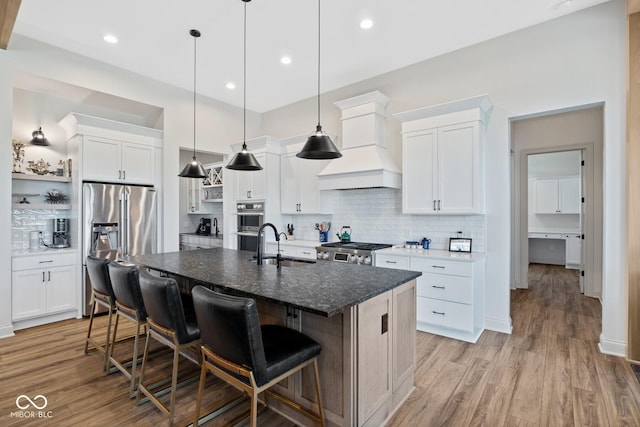 kitchen with a center island with sink, stainless steel appliances, custom range hood, white cabinets, and a kitchen breakfast bar