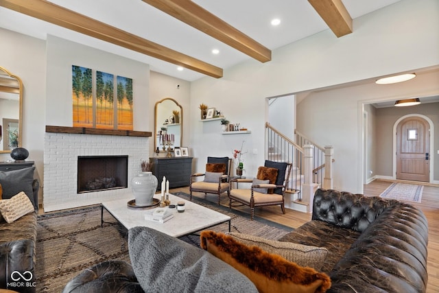 living room with arched walkways, light wood-style floors, beamed ceiling, stairs, and a brick fireplace