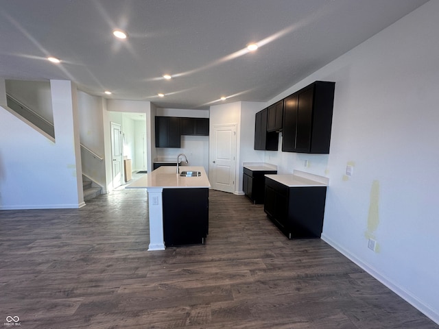 kitchen with a center island with sink, baseboards, dark wood-type flooring, light countertops, and a sink