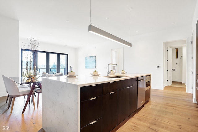 kitchen with sink, dark brown cabinets, a center island with sink, stainless steel dishwasher, and light hardwood / wood-style floors