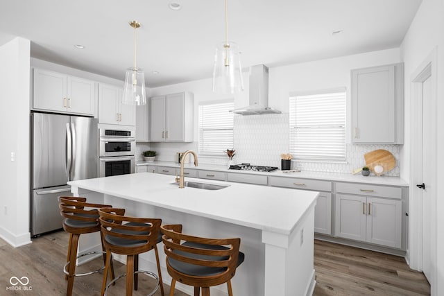 kitchen with pendant lighting, wall chimney exhaust hood, sink, a kitchen island with sink, and stainless steel appliances