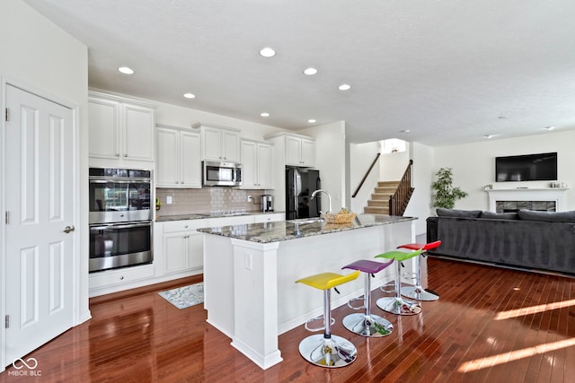 kitchen featuring dark wood-style flooring, decorative backsplash, black appliances, an island with sink, and dark stone countertops