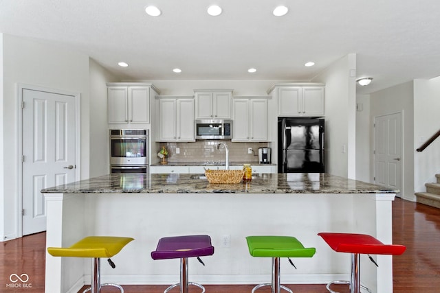 kitchen featuring stainless steel appliances, backsplash, a kitchen island with sink, a sink, and dark stone countertops