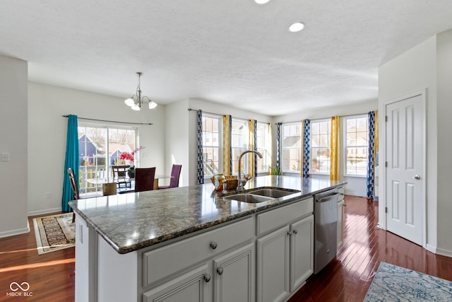 kitchen with dark wood-style flooring, stainless steel dishwasher, a kitchen island with sink, a sink, and dark stone countertops