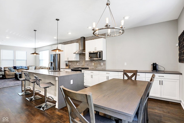 kitchen featuring white cabinets, appliances with stainless steel finishes, wall chimney exhaust hood, decorative light fixtures, and a kitchen island with sink