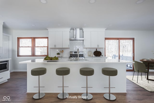 kitchen featuring white cabinetry, an island with sink, a breakfast bar area, and wall chimney range hood