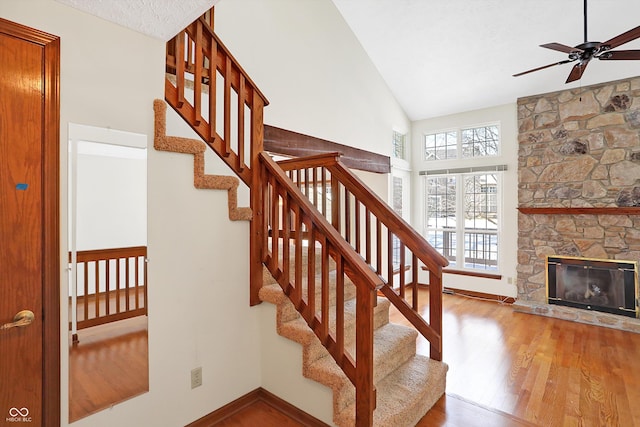 staircase featuring ceiling fan, hardwood / wood-style flooring, high vaulted ceiling, and a stone fireplace