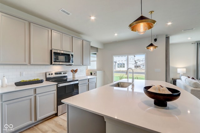 kitchen with hanging light fixtures, appliances with stainless steel finishes, sink, gray cabinets, and an island with sink