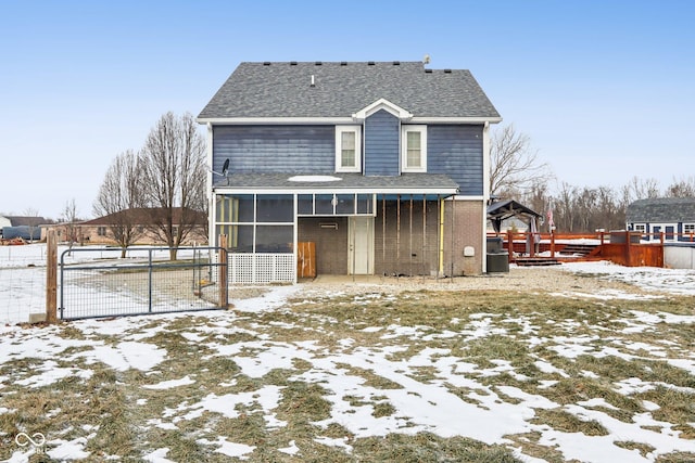 snow covered property featuring a shingled roof, a sunroom, a gate, fence, and brick siding