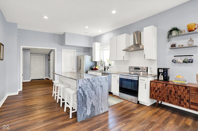 kitchen with appliances with stainless steel finishes, light stone countertops, white cabinets, a kitchen island, and wall chimney exhaust hood