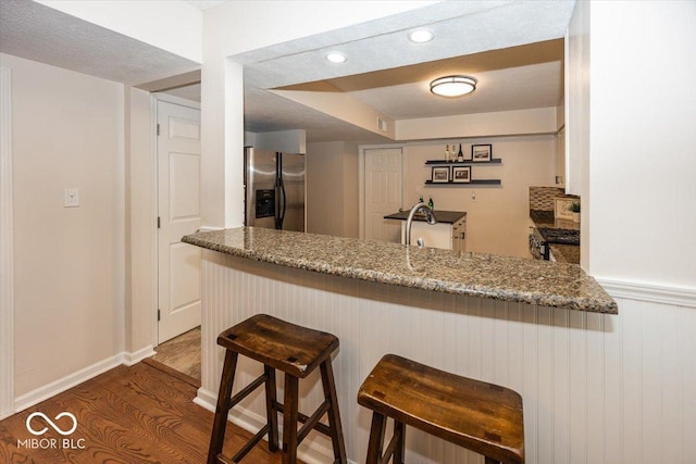 kitchen featuring a kitchen breakfast bar, stainless steel refrigerator with ice dispenser, light stone countertops, dark hardwood / wood-style flooring, and kitchen peninsula