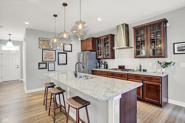 kitchen with sink, wall chimney range hood, backsplash, stainless steel appliances, and an island with sink