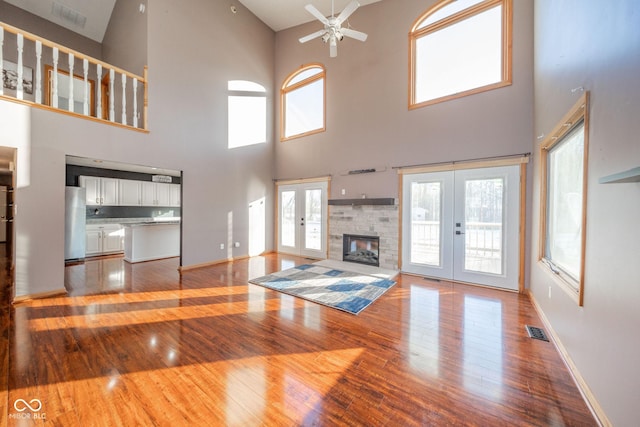 unfurnished living room featuring french doors, a stone fireplace, ceiling fan, a towering ceiling, and hardwood / wood-style floors