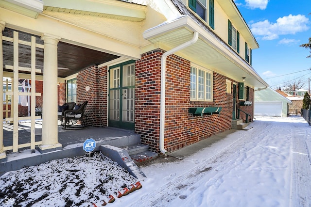 snow covered property featuring covered porch, brick siding, a detached garage, and an outbuilding