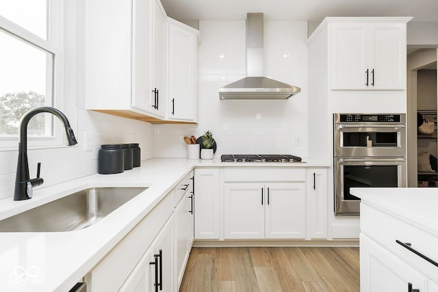 kitchen featuring a sink, gas cooktop, stainless steel double oven, white cabinets, and wall chimney range hood