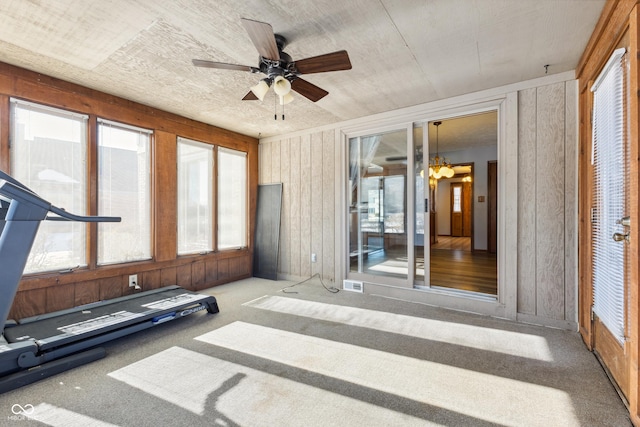 workout area featuring light colored carpet, plenty of natural light, ceiling fan with notable chandelier, and wood walls