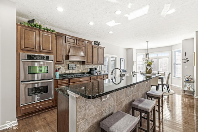 kitchen featuring a breakfast bar, premium range hood, a kitchen island with sink, decorative light fixtures, and stainless steel double oven