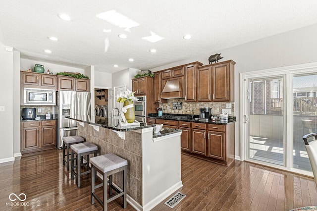 kitchen with extractor fan, appliances with stainless steel finishes, an island with sink, dark stone countertops, and a kitchen breakfast bar