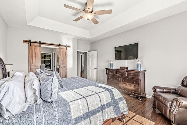 bedroom with a raised ceiling, a barn door, dark hardwood / wood-style floors, and ceiling fan