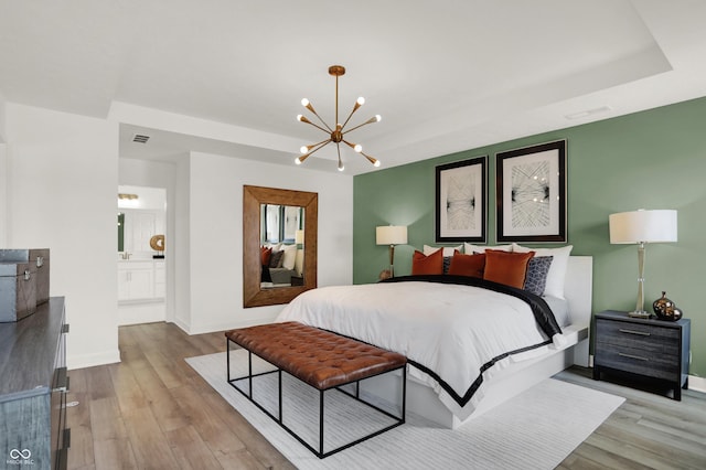 bedroom featuring an inviting chandelier, connected bathroom, a tray ceiling, and light wood-type flooring