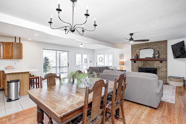 dining space featuring light hardwood / wood-style flooring, ceiling fan with notable chandelier, a fireplace, and a textured ceiling