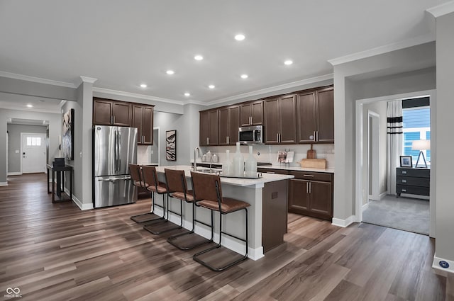 kitchen featuring dark wood-type flooring, dark brown cabinets, a center island with sink, appliances with stainless steel finishes, and a kitchen breakfast bar