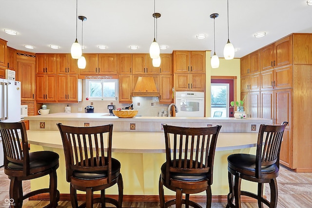 kitchen featuring pendant lighting, backsplash, a kitchen breakfast bar, custom exhaust hood, and white appliances