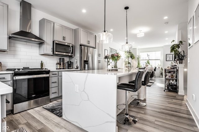 kitchen featuring gray cabinetry, stainless steel appliances, a kitchen island with sink, and wall chimney range hood