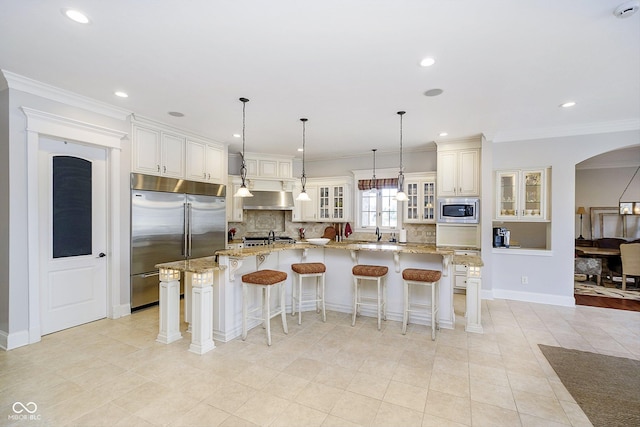 kitchen featuring light stone counters, built in appliances, a large island with sink, pendant lighting, and exhaust hood