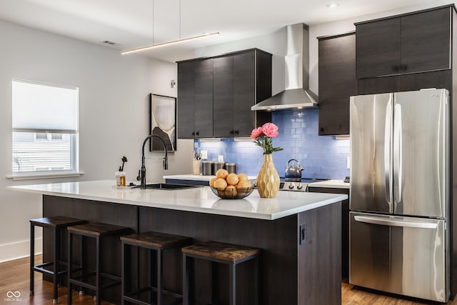 kitchen with an island with sink, sink, stainless steel refrigerator, and wall chimney exhaust hood