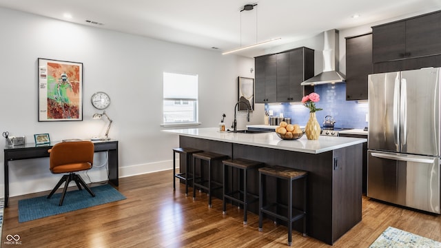 kitchen with a breakfast bar, pendant lighting, stainless steel fridge, a center island with sink, and wall chimney exhaust hood