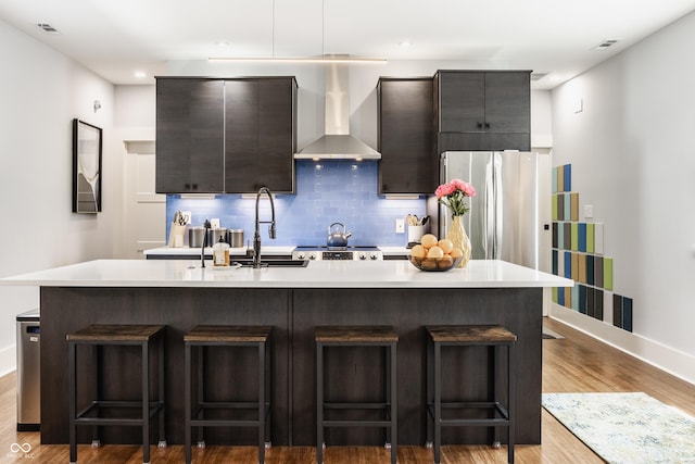 kitchen with stainless steel refrigerator, wall chimney exhaust hood, an island with sink, and a breakfast bar area