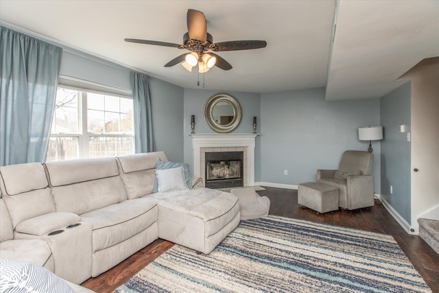 living room featuring a tiled fireplace, ceiling fan, baseboards, and dark wood-style flooring