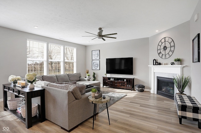 living room with a tiled fireplace, ceiling fan, a textured ceiling, and light wood-type flooring