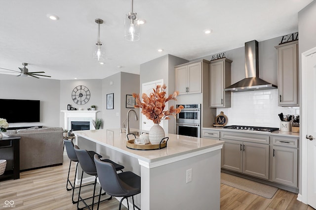 kitchen with gray cabinets, appliances with stainless steel finishes, decorative light fixtures, a kitchen island with sink, and wall chimney range hood