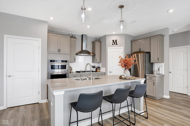 kitchen with appliances with stainless steel finishes, wall chimney exhaust hood, decorative light fixtures, and a kitchen island with sink