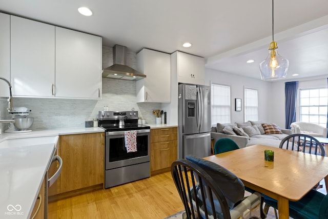 kitchen featuring appliances with stainless steel finishes, white cabinetry, hanging light fixtures, light hardwood / wood-style floors, and wall chimney exhaust hood