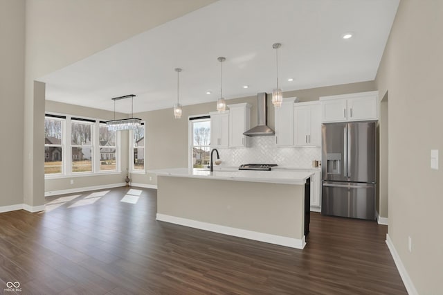 kitchen with wall chimney exhaust hood, white cabinetry, decorative light fixtures, appliances with stainless steel finishes, and a kitchen island with sink