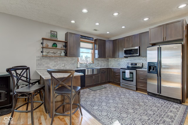 kitchen featuring a peninsula, decorative backsplash, dark brown cabinets, light wood-style floors, and appliances with stainless steel finishes