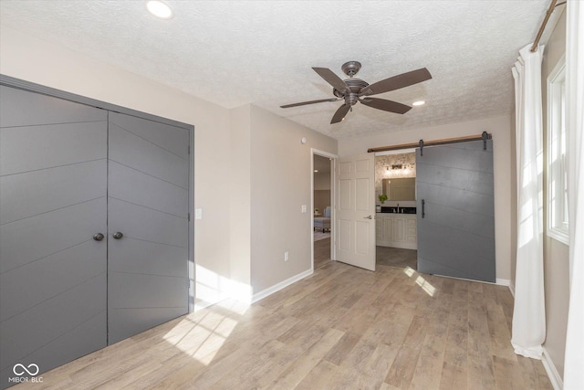 unfurnished bedroom with connected bathroom, a textured ceiling, light wood-type flooring, ceiling fan, and a barn door