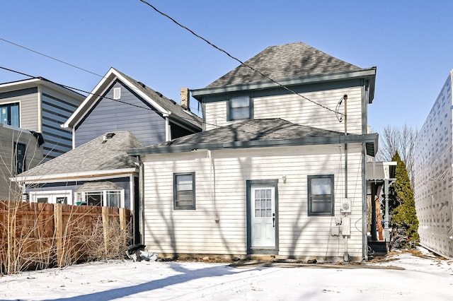 snow covered rear of property with a shingled roof and fence