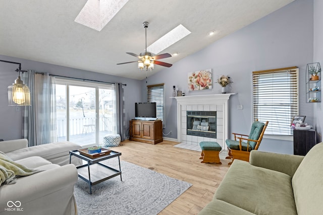 living room featuring light hardwood / wood-style flooring, a tile fireplace, ceiling fan, vaulted ceiling with skylight, and a textured ceiling