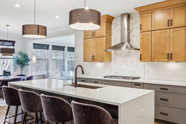 kitchen with sink, hanging light fixtures, stainless steel gas cooktop, a center island with sink, and wall chimney exhaust hood