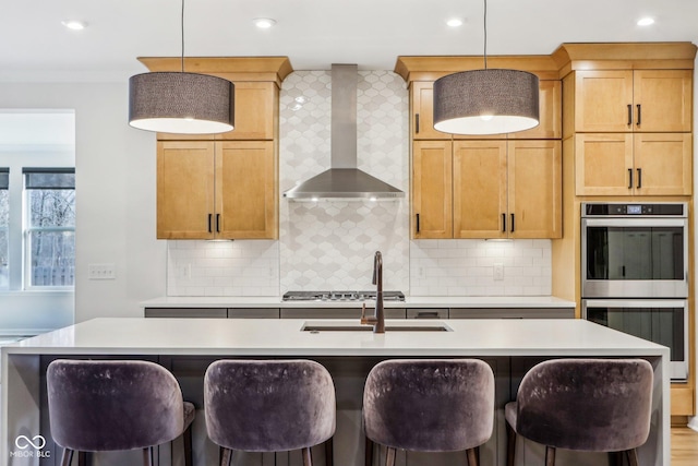 kitchen with wall chimney range hood, decorative light fixtures, stainless steel appliances, and an island with sink