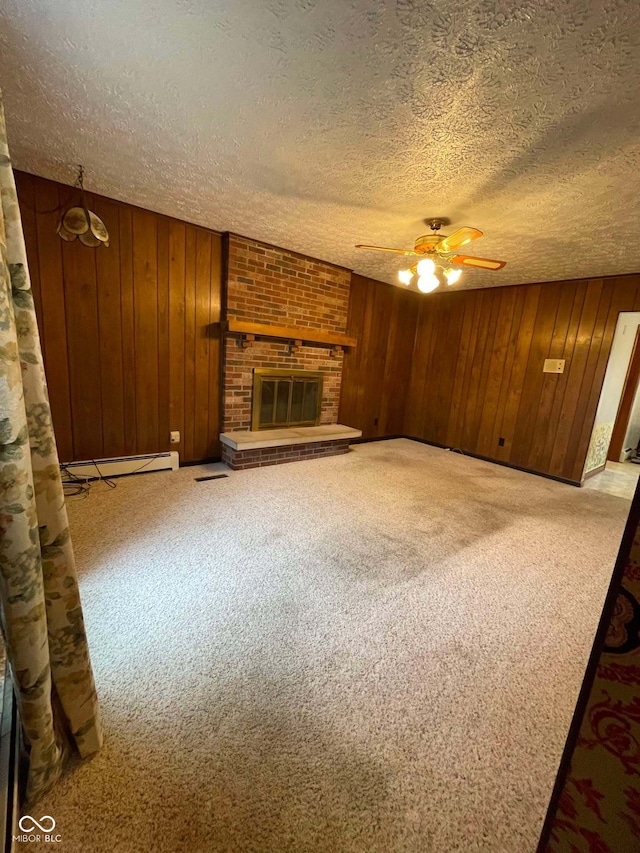 unfurnished living room featuring wooden walls, carpet floors, baseboard heating, a brick fireplace, and a textured ceiling