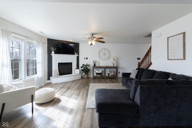 living room featuring visible vents, ceiling fan, wood finished floors, stairs, and a brick fireplace