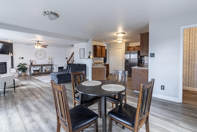 dining area featuring a fireplace with raised hearth, wood finished floors, a ceiling fan, baseboards, and stairs