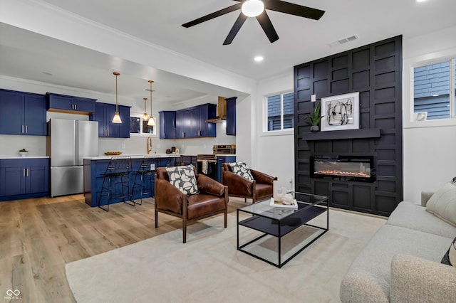 living room featuring a large fireplace, sink, ceiling fan, crown molding, and light wood-type flooring