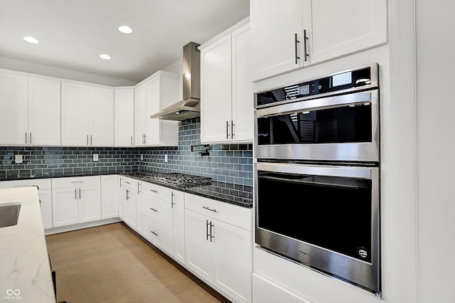 kitchen with wall chimney exhaust hood, light stone counters, white cabinetry, stainless steel appliances, and light hardwood / wood-style floors