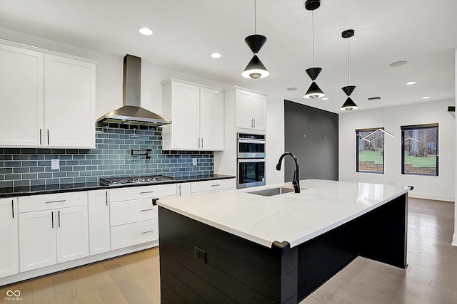 kitchen featuring sink, decorative light fixtures, an island with sink, stainless steel appliances, and wall chimney range hood